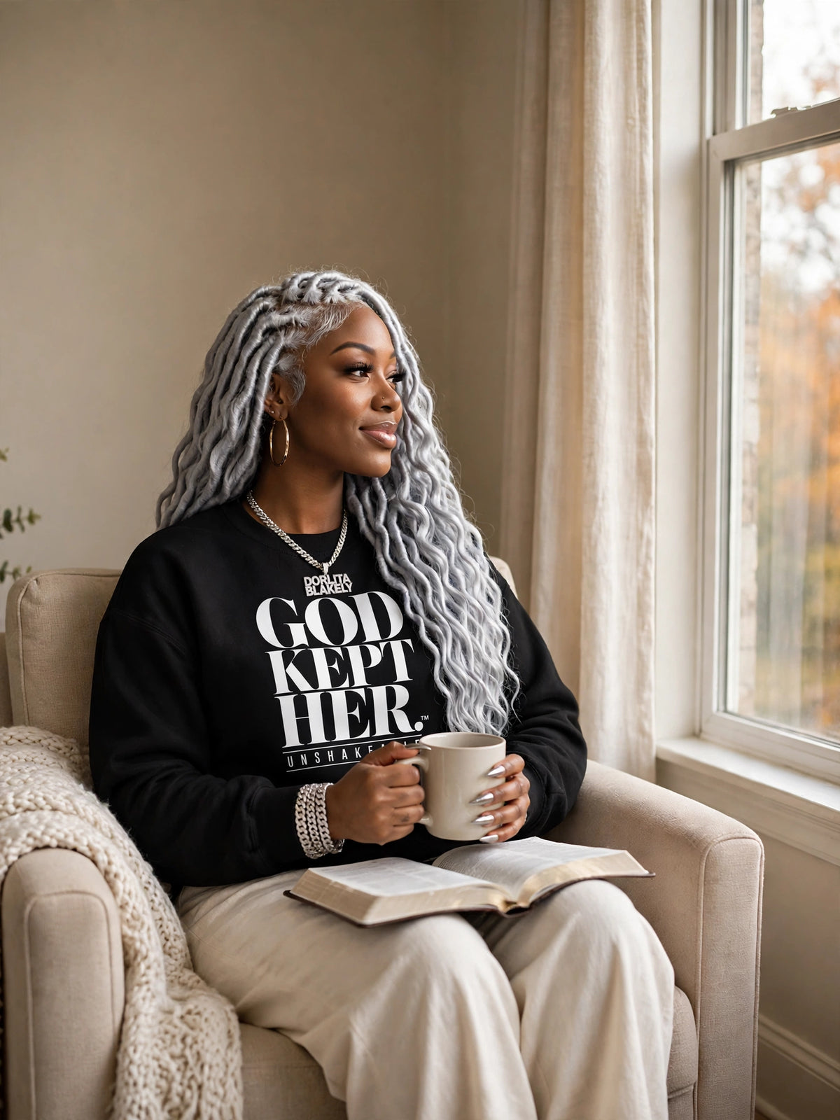 Woman wearing the black God Kept Her sweatshirt by a window with coffee and Bible